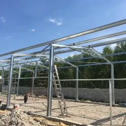 Stalowa konstrukcja hali w trakcie montażu, widoczne rusztowania, drabiny i przygotowane podłoże z gruzem, na tle zieleni i błękitnego nieba.