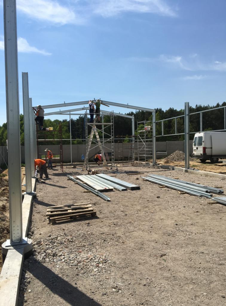 Montaż konstrukcji stalowej hali: pracownicy na rusztowaniach łączą elementy szkieletu, widoczne słupy i belki, w tle las i biały bus.