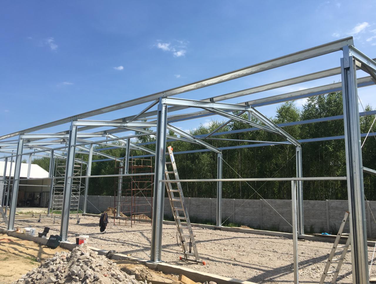 Stalowa konstrukcja hali w trakcie montażu, widoczne rusztowania, drabiny i przygotowane podłoże z gruzem, na tle zieleni i błękitnego nieba.