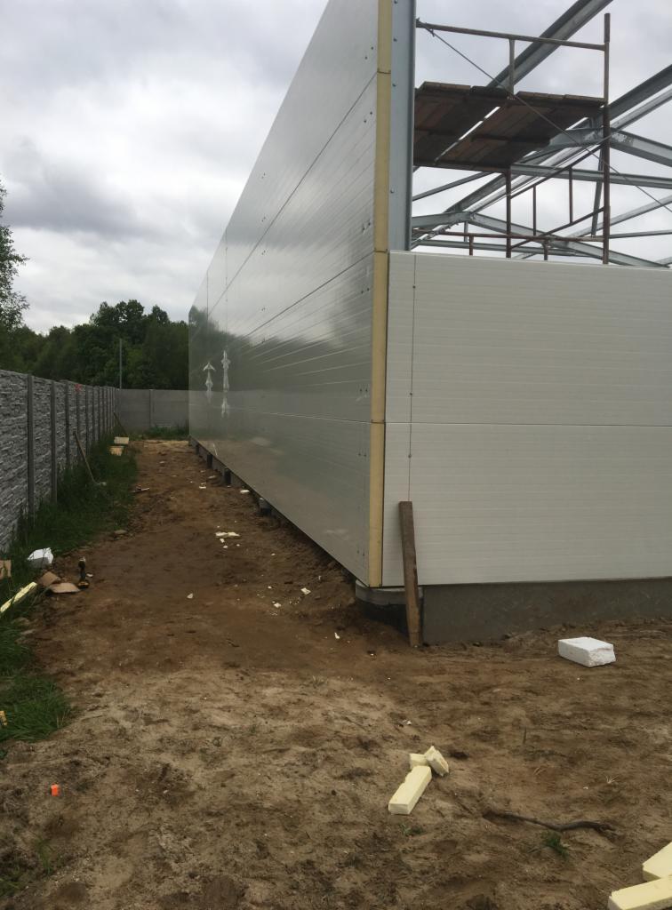 Montaż szarych i białych płyt warstwowych na konstrukcji stalowej budynku, widoczny fragment rusztowania i grunt.