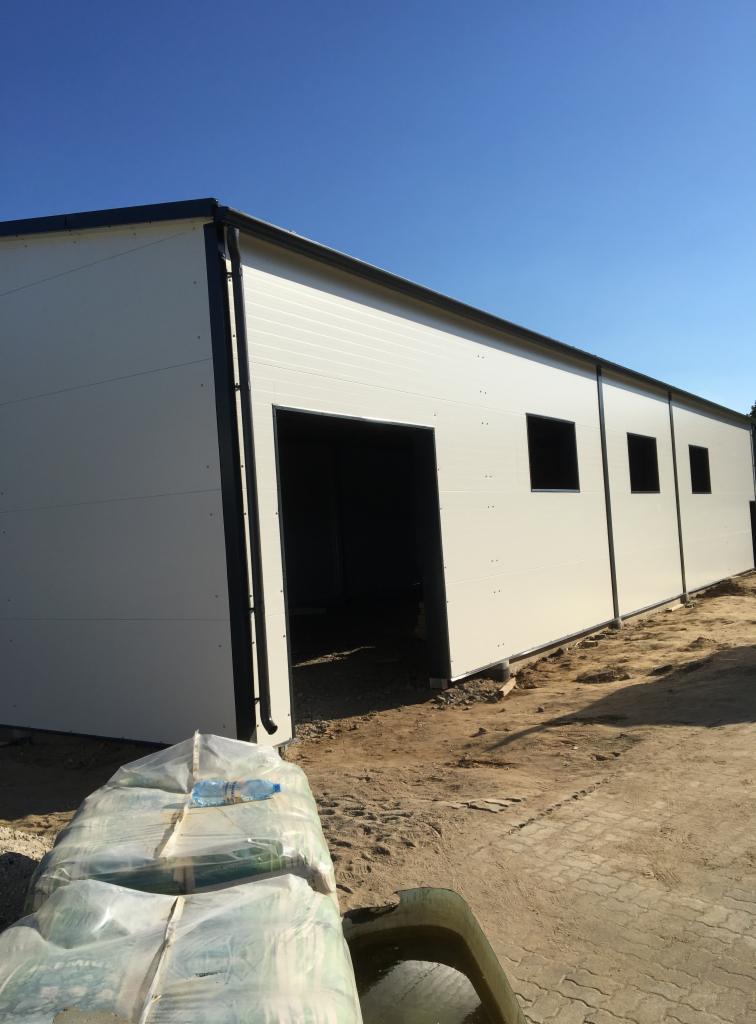 Budynek w trakcie budowy, wykonany z białych płyt warstwowych z czarnymi obramowaniami, widoczne otwory okienne i drzwiowe, teren wokół budynku częściowo utwardzony, z przodu leżą materiały...