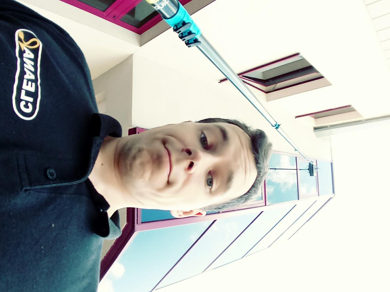 Mężczyzna robiący selfie z kijem teleskopowym do mycia okien, widoczne okna budynku odbijające niebo.