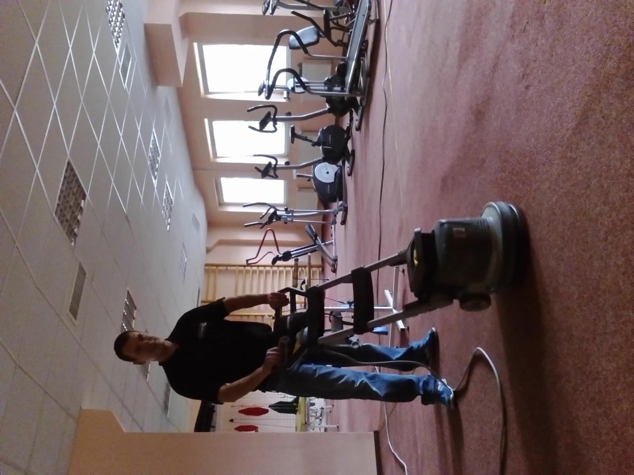 Pracownik w czarnej koszulce i jeansach obsługuje maszynę do czyszczenia dywanów w siłowni, w tle sprzęt do ćwiczeń i okna.