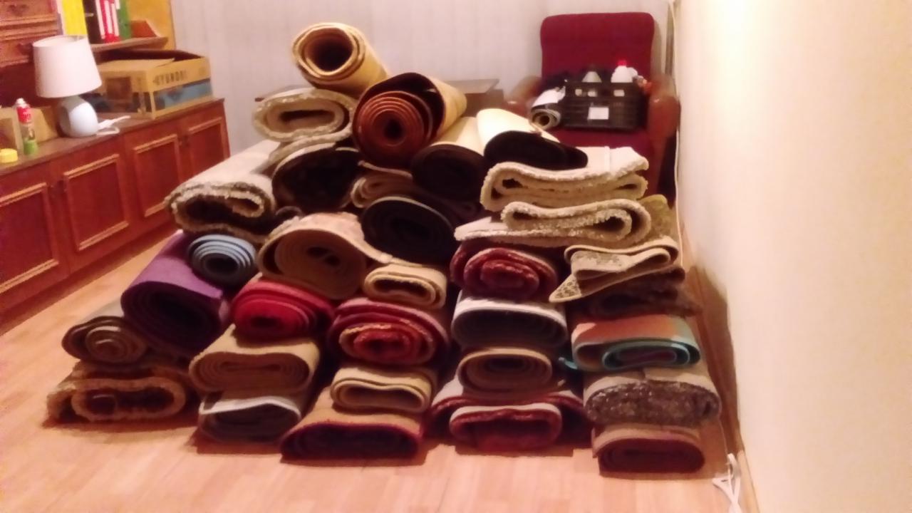 Stos zwiniętych dywanów i wykładzin w różnych kolorach i wzorach, ułożonych na drewnianej podłodze w pomieszczeniu z meblami w tle.