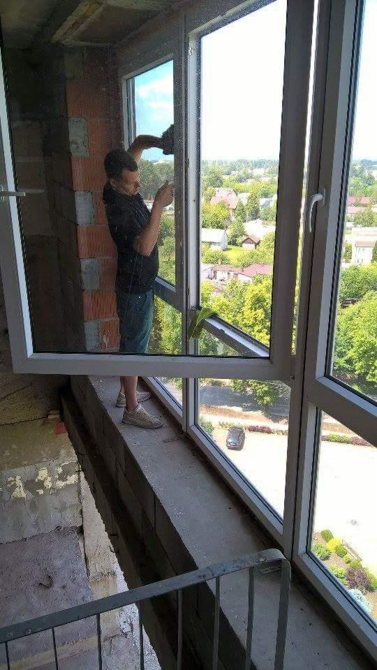 Mężczyzna w trakcie montażu okna balkonowego z widokiem na osiedle domów jednorodzinnych, widoczne surowe wykończenie balkonu z cegłą i betonem.