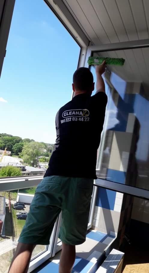 Mężczyzna w czarnej koszulce z logo firmy sprzątającej CLEANAZ i numerem telefonu 722 03 44 22 myje okno z zewnątrz za pomocą myjki z zieloną nakładką, widok z balkonu na miasto w słoneczny dzień.