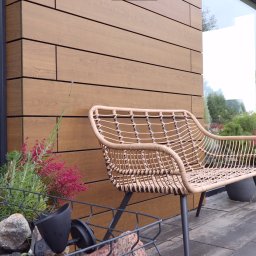 Dehha Szymon Radko - Beżowy, pleciony fotel ogrodowy stoi na szarym bruku przed budynkiem z elewacją z paneli imitujących drewno. Obok widoczna skrzynka z kamieniami i roślinami oraz duże okno.