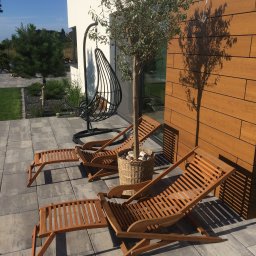 Dehha Szymon Radko - Dwa drewniane leżaki z podnóżkami na szarym bruku, obok ściany wyłożonej drewnianymi panelami i drzewka w wiklinowym koszu, w tle wiszące krzesło.