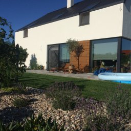 Dehha Szymon Radko - Nowoczesny dom jednorodzinny z dużymi oknami, drewnianą elewacją i tarasem z leżakami, otoczony ogrodem z kamieniami, lawendą i basenem dla dzieci.