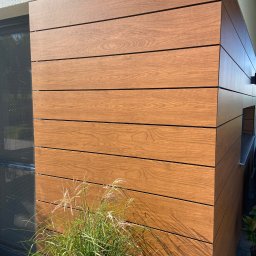 Dehha Szymon Radko - Drewniana elewacja budynku z poziomych paneli, oświetlona słońcem, z ozdobną trawą u podstawy i widocznym fragmentem okna.