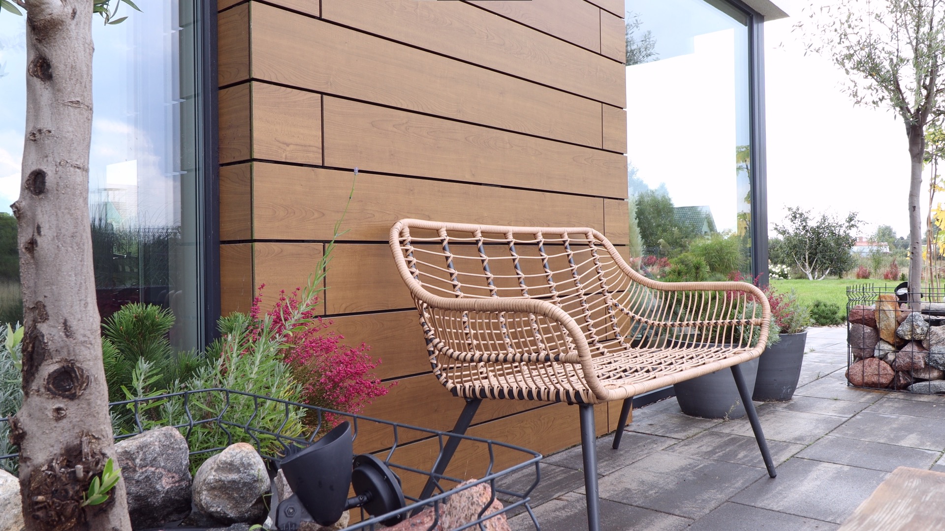 Beżowy, pleciony fotel ogrodowy stoi na szarym bruku przed budynkiem z elewacją z paneli imitujących drewno. Obok widoczna skrzynka z kamieniami i roślinami oraz duże okno.