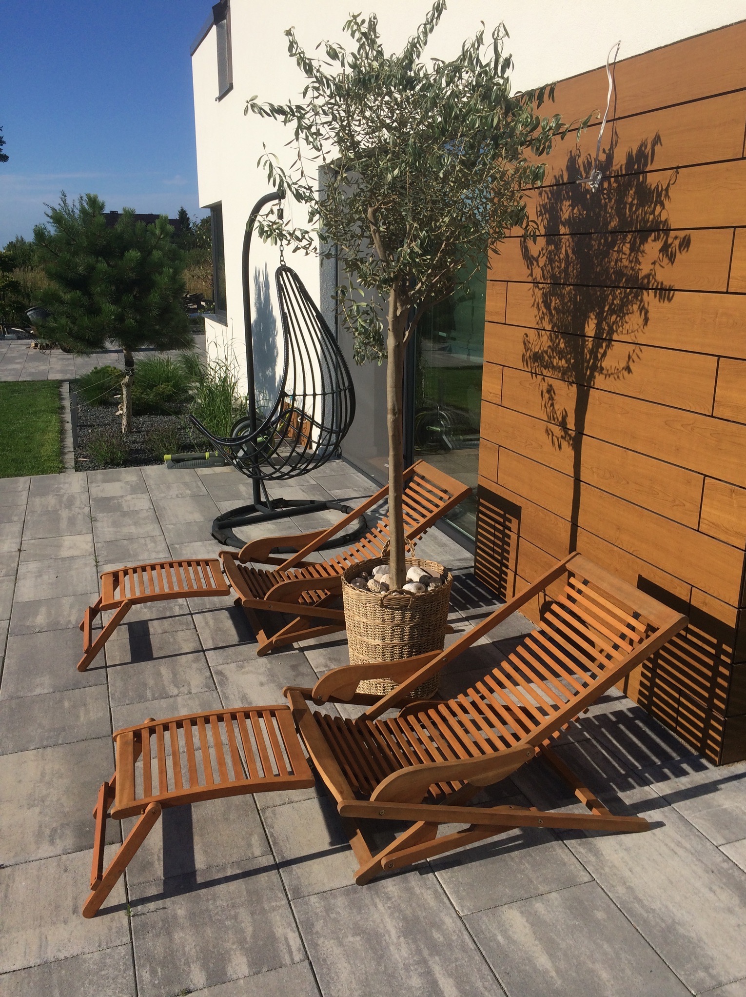 Dwa drewniane leżaki z podnóżkami na szarym bruku, obok ściany wyłożonej drewnianymi panelami i drzewka w wiklinowym koszu, w tle wiszące krzesło.