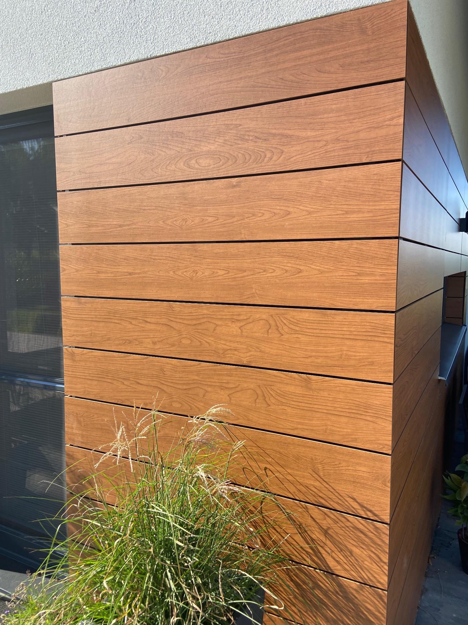 Drewniana elewacja budynku z poziomych paneli, oświetlona słońcem, z ozdobną trawą u podstawy i widocznym fragmentem okna.