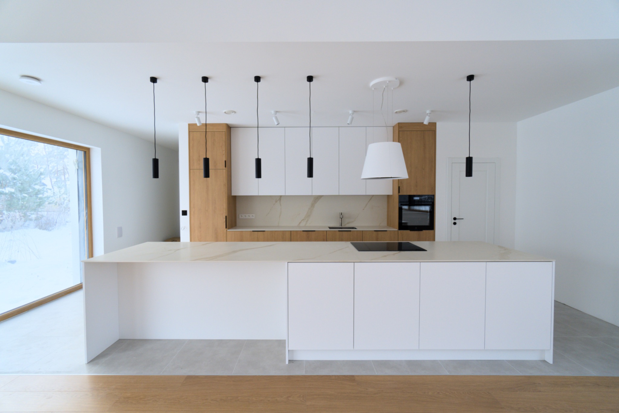 Nowoczesna kuchnia z wyspą, białe fronty szafek dolnych i górnych, drewniane elementy, marmurowy blat, minimalistyczne czarne lampy wiszące, duże okno z widokiem na zimowy krajobraz.
