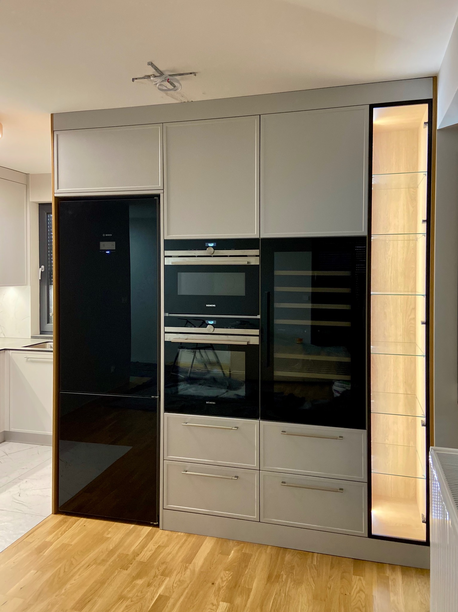 Szara zabudowa kuchenna z wbudowanym sprzętem AGD: lodówka Bosch z czarnym frontem, piekarnik i mikrofalówka Siemens, chłodziarka do wina oraz przeszklona witryna z podświetleniem, na drewnianej...