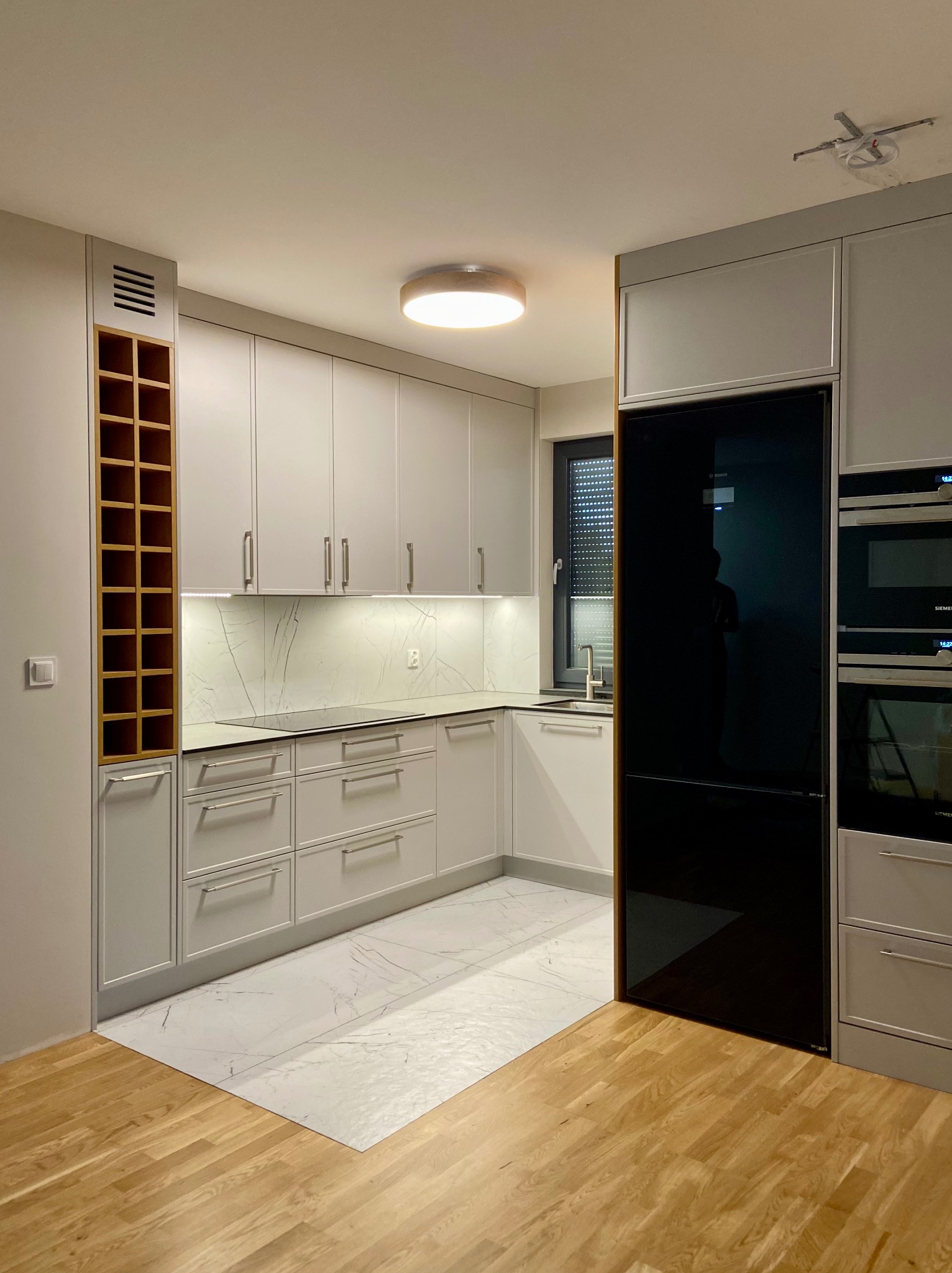 Nowoczesna kuchnia w odcieniach szarości z drewnianymi akcentami, marmurowym blatem i podświetleniem LED, widoczna zabudowa z piekarnikiem i lodówką side-by-side.