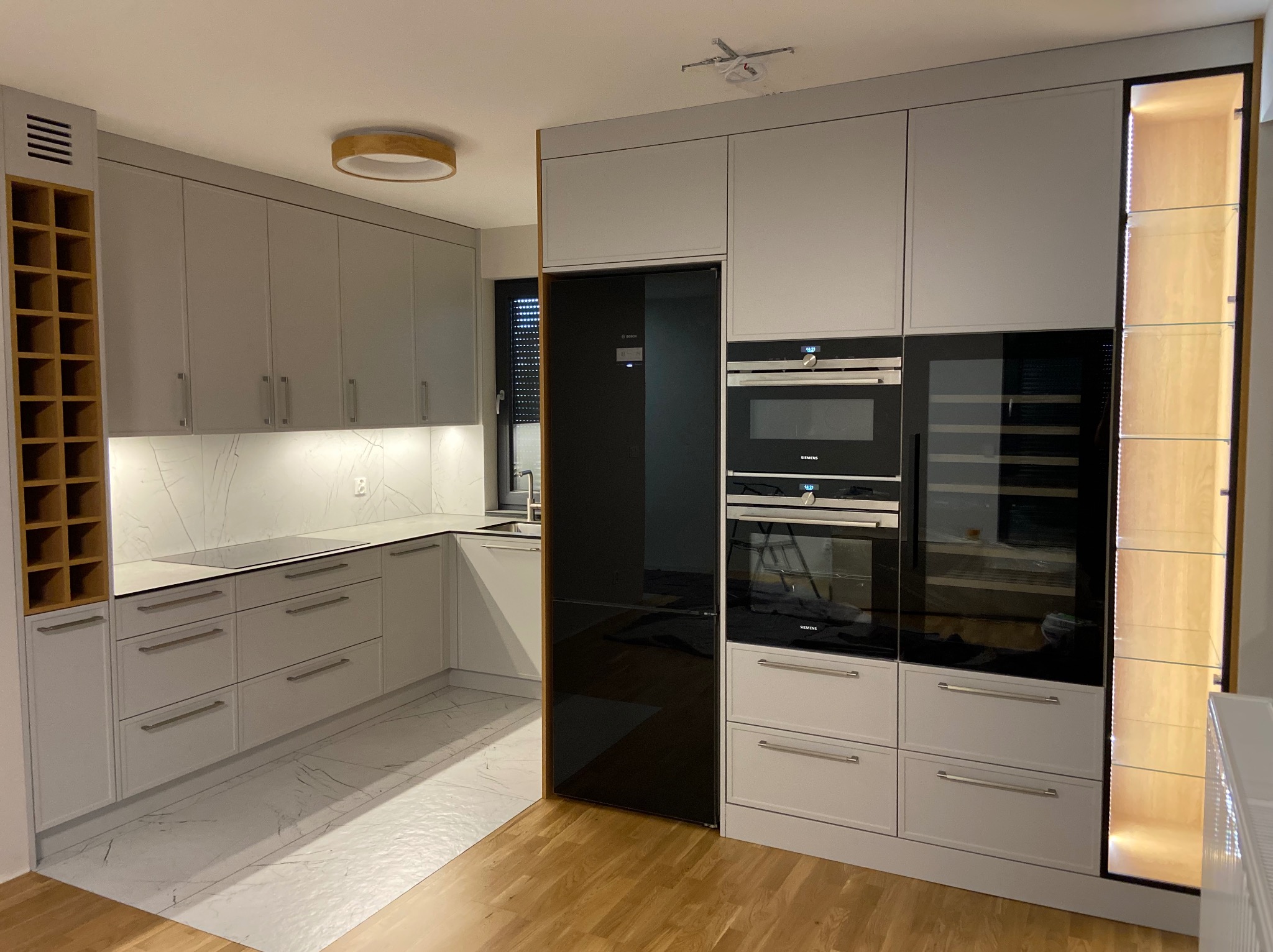 Nowoczesna kuchnia w odcieniach szarości z marmurowym blatem i podłogą, zabudowana lodówka i sprzęt AGD marki Siemens, drewniany regał na wino i oświetlenie podszafkowe.