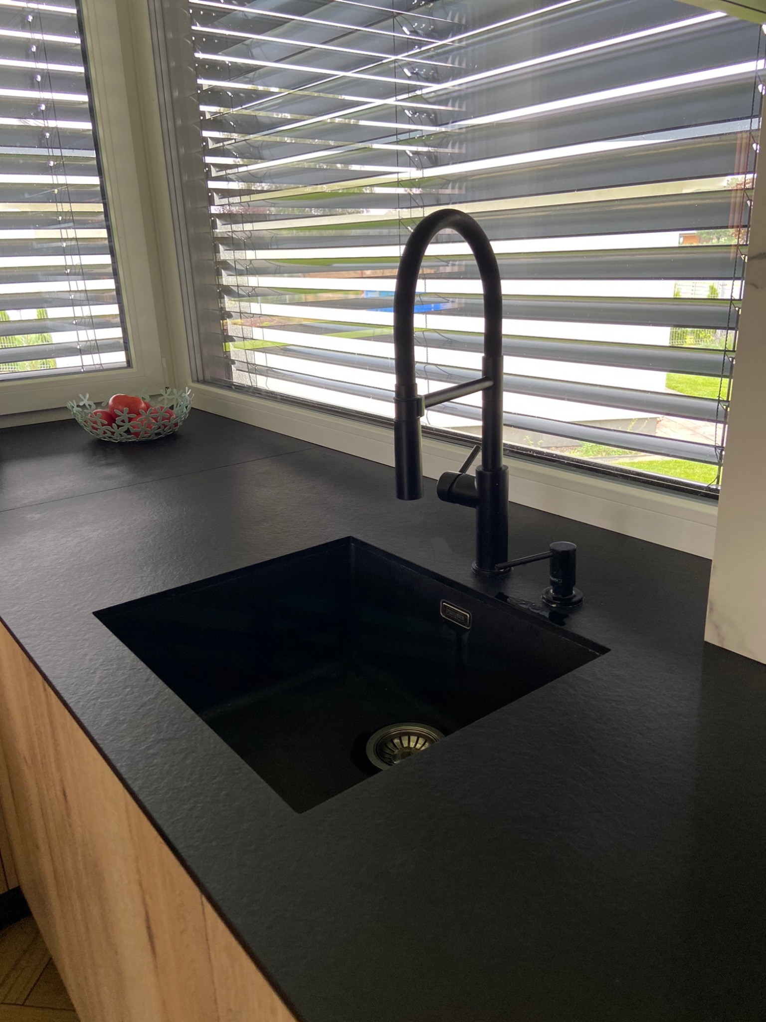 Nowoczesna kuchnia: czarny, kamienny blat z wbudowanym zlewozmywakiem i czarną, minimalistyczną baterią, widok na okno z żaluzjami.
