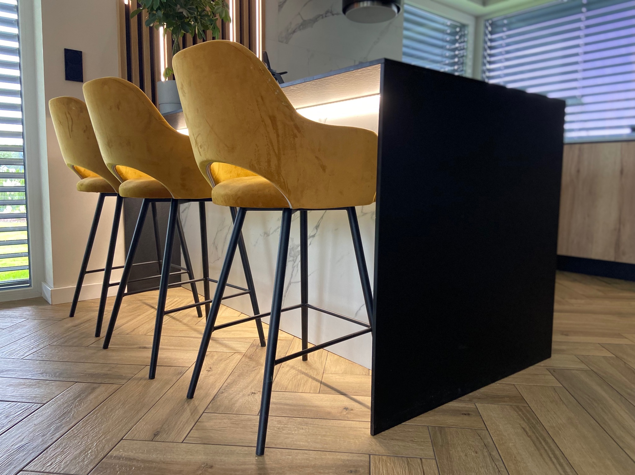 Trzy welurowe hokery w odcieniach złota z czarnymi nogami ustawione przy czarnym blacie wyspy kuchennej z podświetleniem LED, na podłodze parkiet ułożony w jodełkę.
