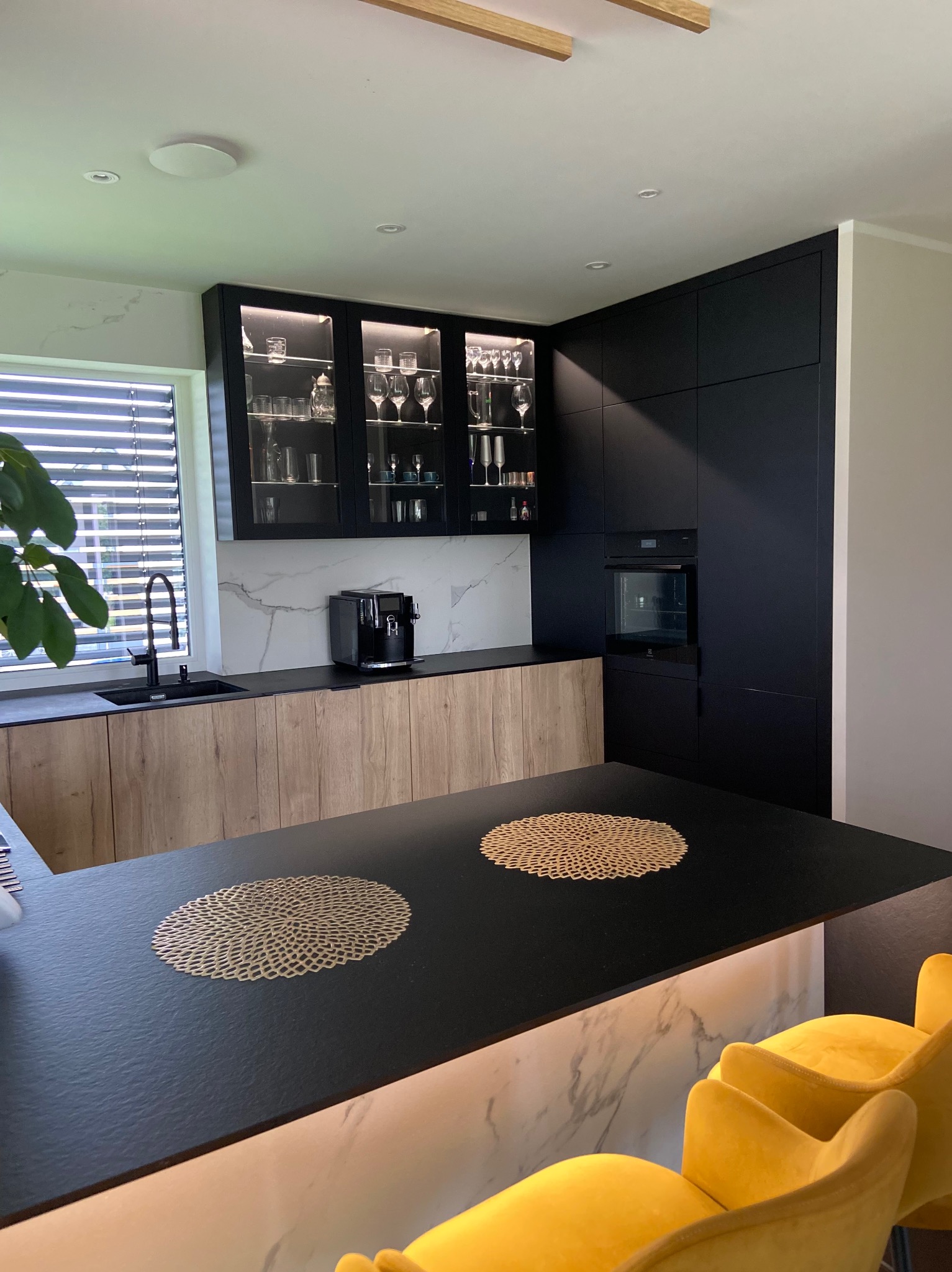 Nowoczesna kuchnia z czarnymi szafkami wiszącymi z podświetleniem i szklanymi frontami, jasnymi drewnianymi szafkami dolnymi, czarnym blatem, marmurowym backsplashiem i wyspą z czarnym blatem,...