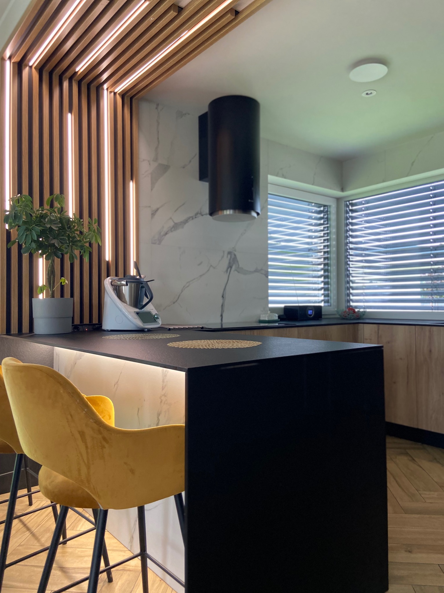 Nowoczesna kuchnia z czarnym blatem, złotymi hokerami i dekoracyjną ścianą z drewnianych listew z podświetleniem LED. Widoczny sprzęt AGD i marmurowe wykończenie ściany.