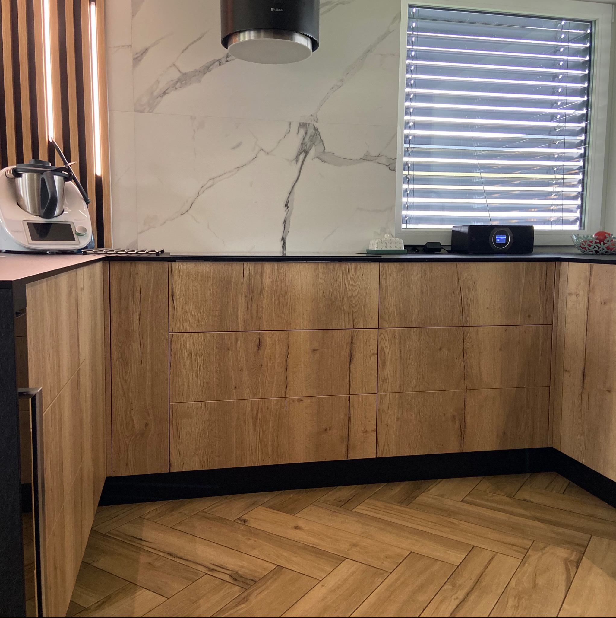 Fronty szafek kuchennych z drewnopodobnym wzorem, czarny blat, biała marmurowa ściana, podłoga ułożona w jodełkę, widoczny robot kuchenny i radio.