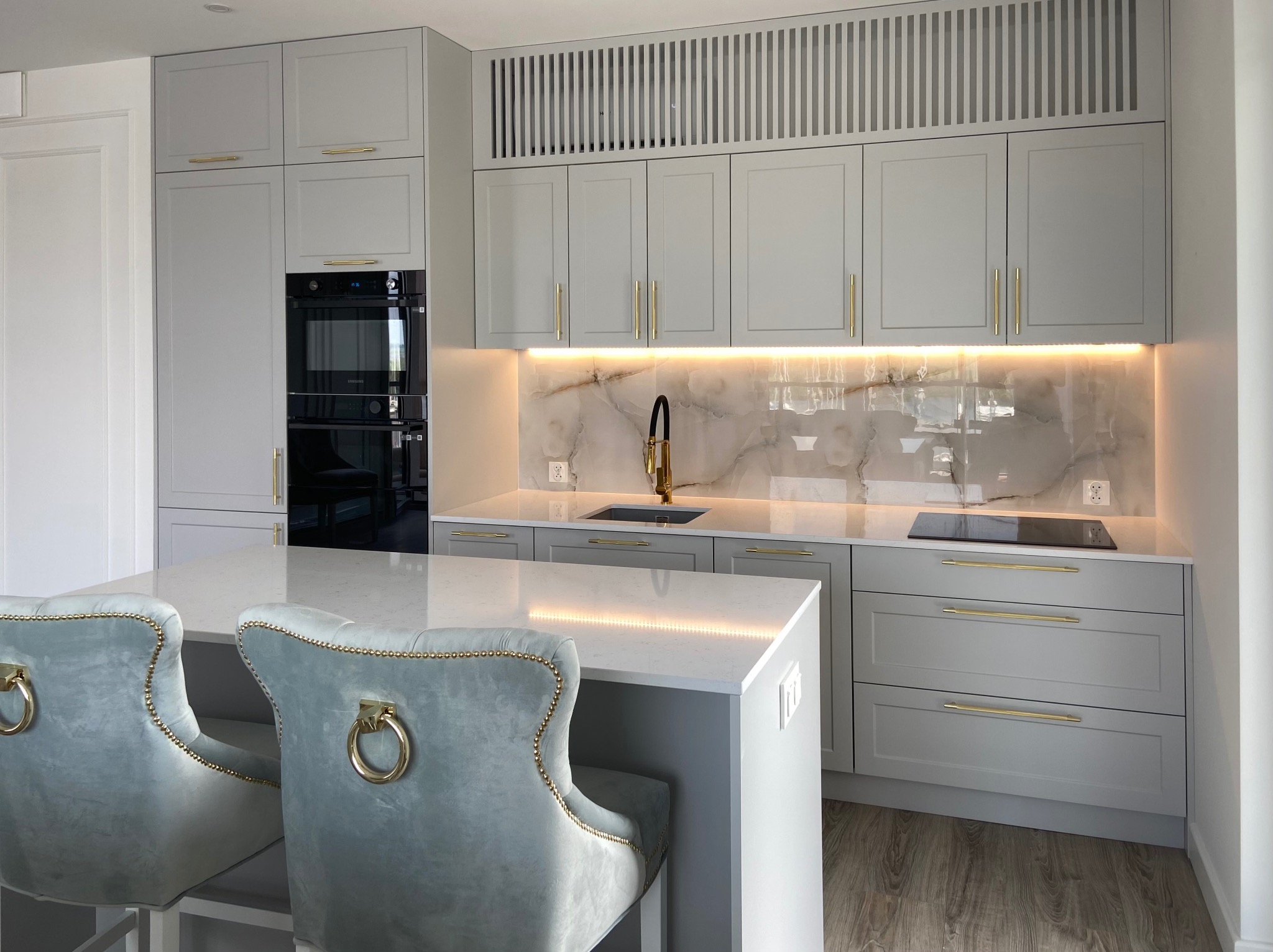Nowoczesna kuchnia w odcieniach szarości z białym blatem i złotymi akcentami, oświetlenie LED pod szafkami, eleganckie krzesła barowe z welurowym obiciem i złotymi uchwytami.