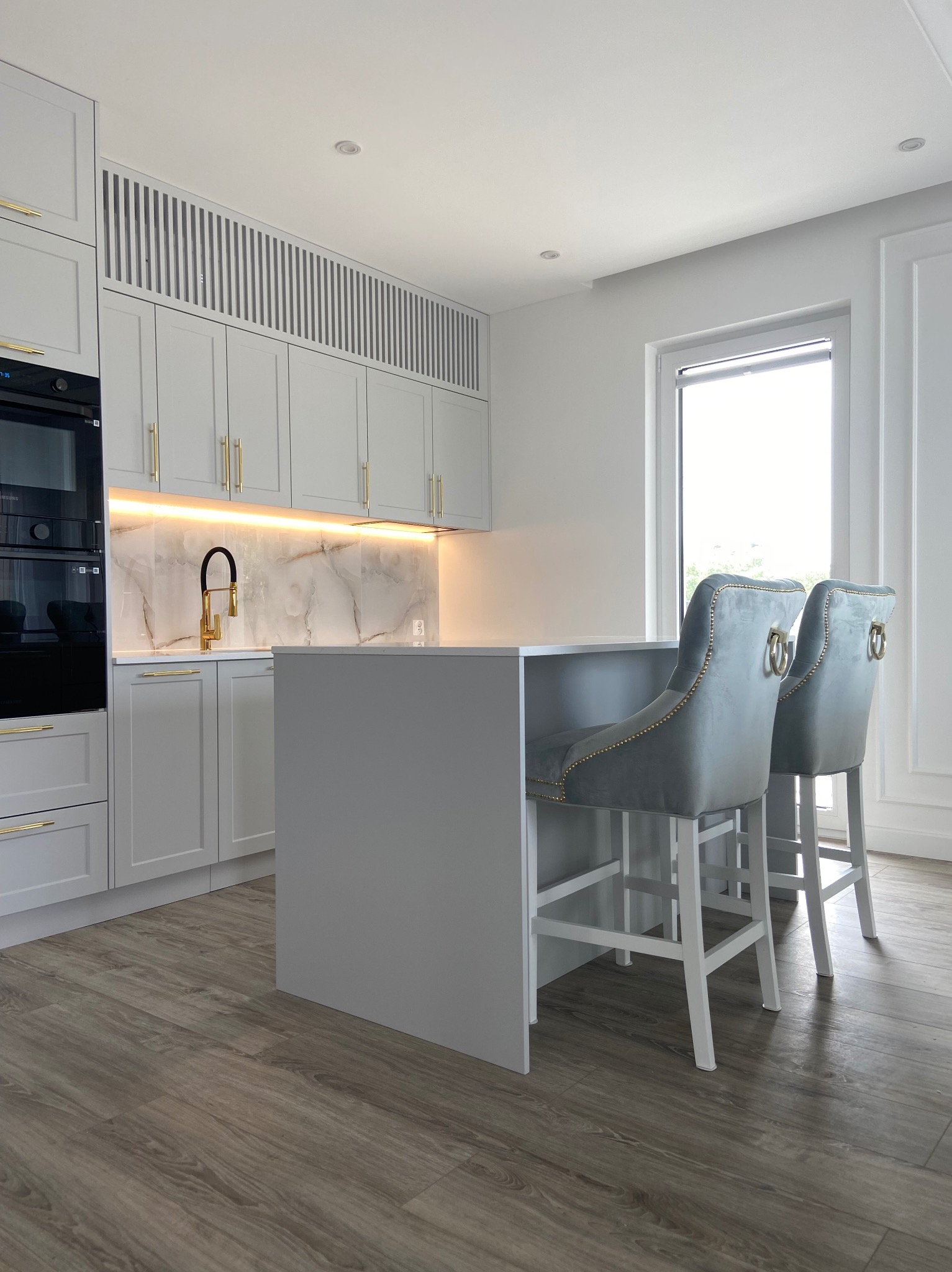 Jasnoszara kuchnia z wyspą, złotą armaturą i marmurowym backsplashiem podświetlonym LED, uzupełniona dwoma szarymi hokerami z dekoracyjnymi złotymi elementami.
