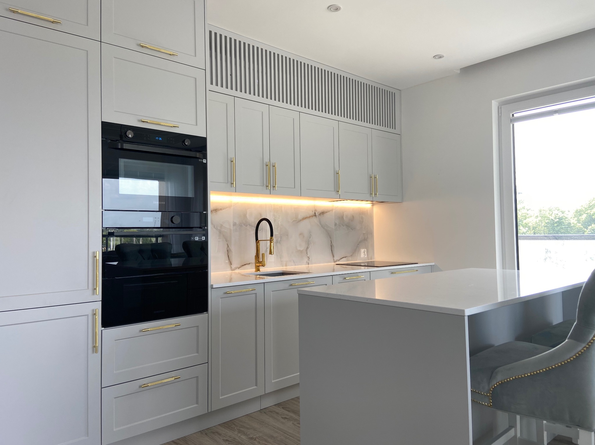 Nowoczesna kuchnia w odcieniach szarości z złotymi uchwytami, marmurowym backsplashiem i wyspą kuchenną z szarym krzesłem barowym.