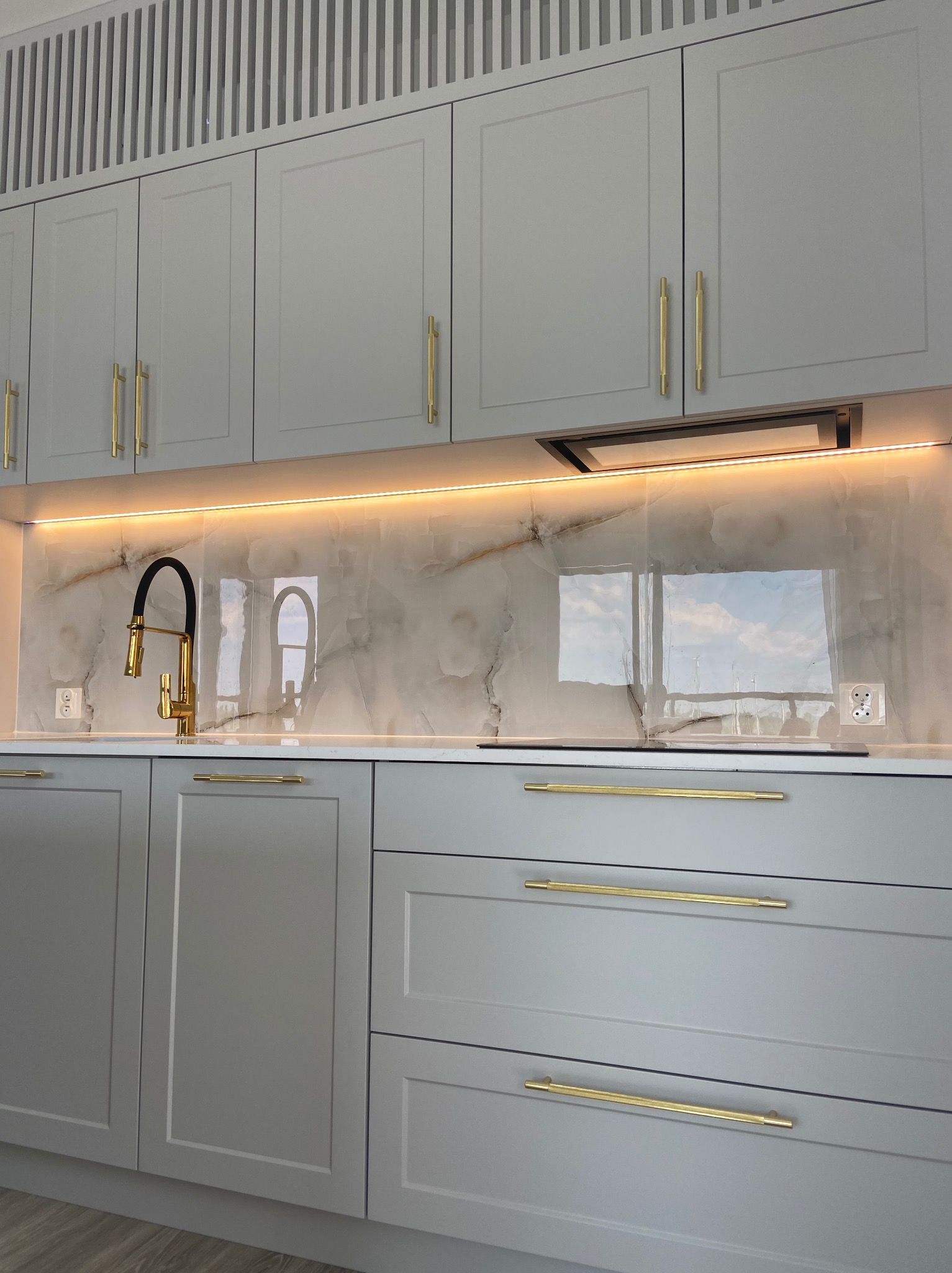 Szare szafki kuchenne z złotymi uchwytami, marmurowy panel nad blatem podświetlony ledami, czarno-złota bateria kuchenna, widok na okno odbijający się w panelu.
