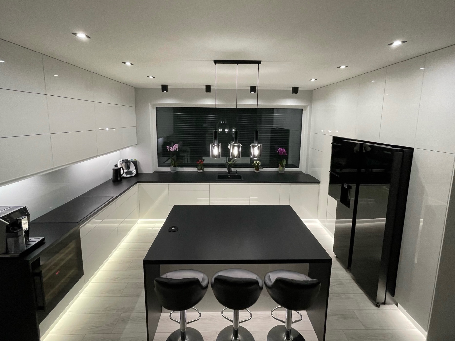 Nowoczesna kuchnia z białymi, błyszczącymi szafkami, czarnym blatem i wyspą z trzema hokerami, oświetlona ledami pod szafkami i wiszącymi lampami nad wyspą.