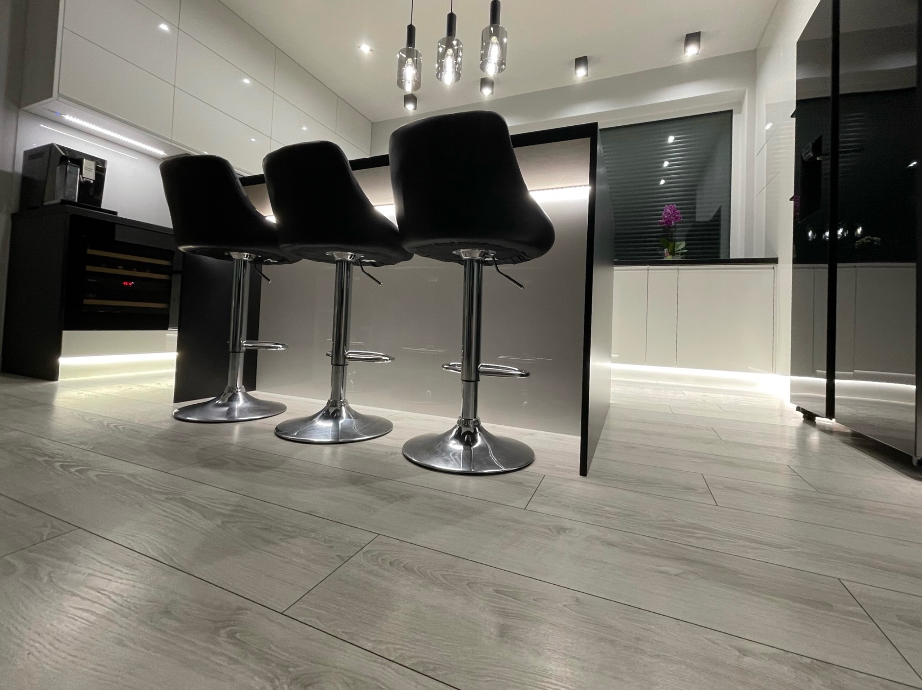 Nowoczesna, minimalistyczna kuchnia z białymi frontami, czarnym barkiem i trzema hokerami z czarnymi siedziskami na chromowanych nogach. Oświetlenie LED pod szafkami i wiszące lampy nad barkiem.