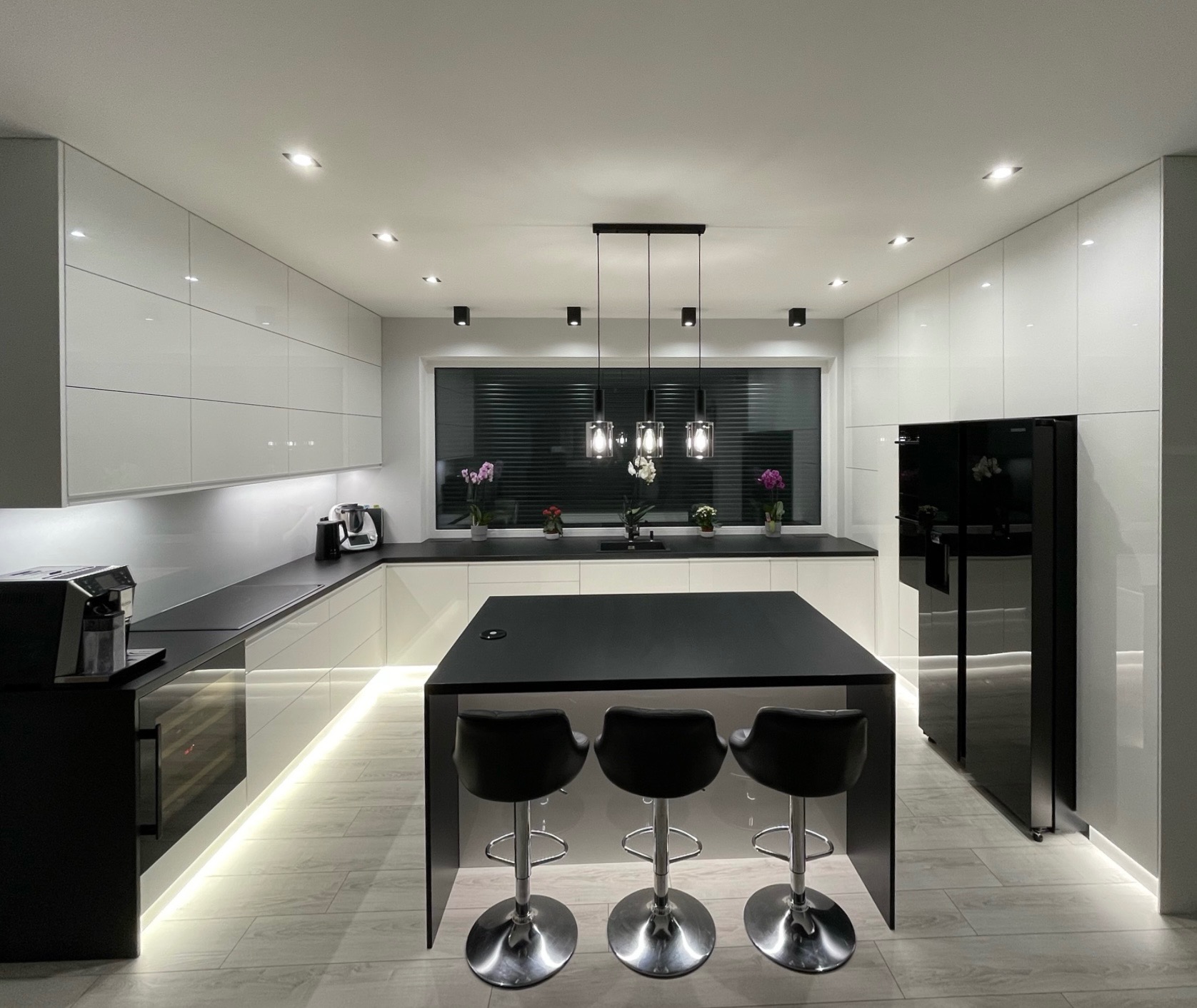 Nowoczesna kuchnia z białymi, lakierowanymi frontami szafek, czarnym blatem i wyspą z trzema hokerami, podświetleniem LED pod szafkami i oknem z żaluzjami.
