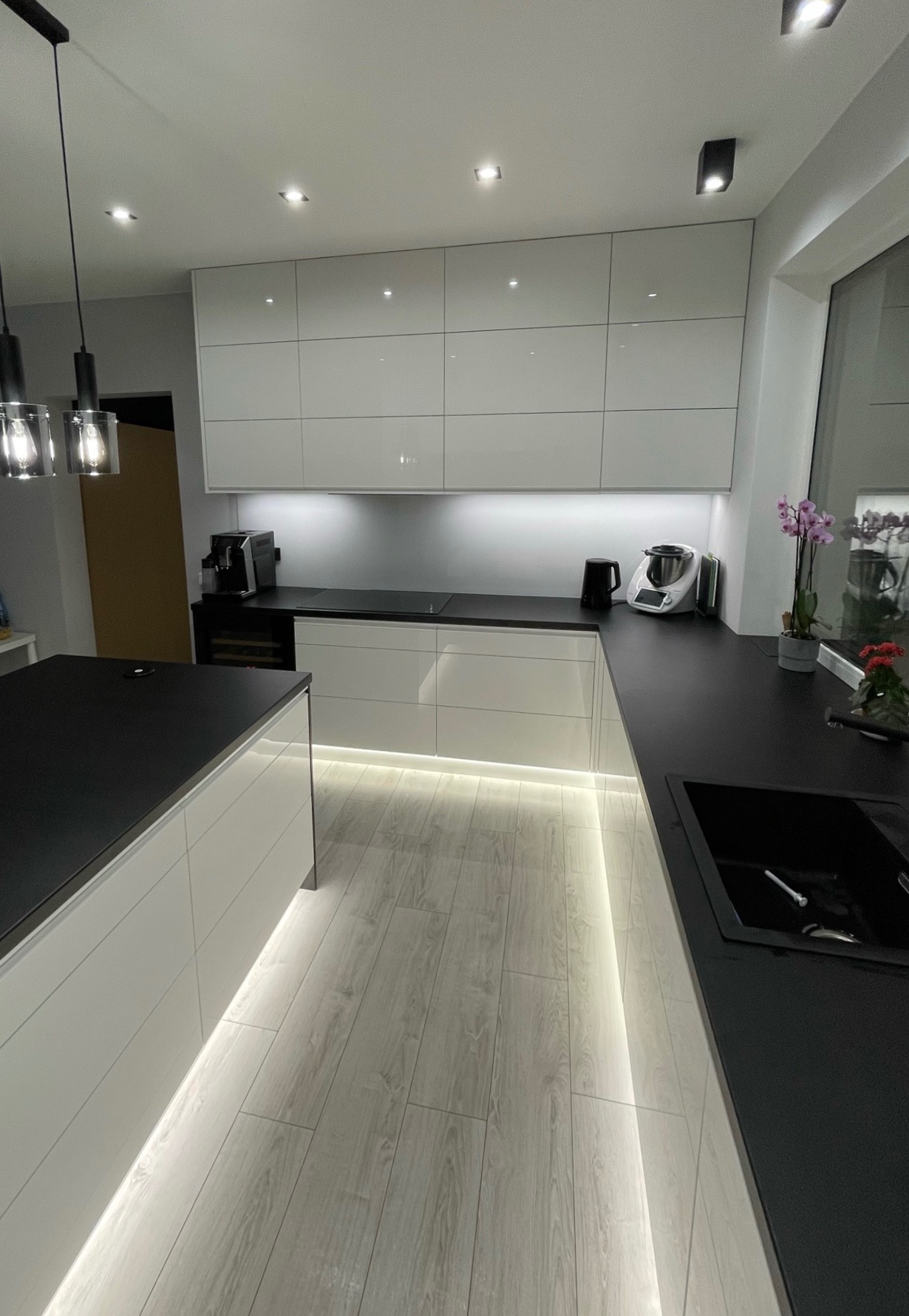 Nowoczesna kuchnia: białe, błyszczące szafki wiszące i dolne z podświetleniem LED przy podłodze, czarny blat, wyspa kuchenna, widoczne sprzęty AGD (ekspres do kawy, robot kuchenny), drewnopodobna...