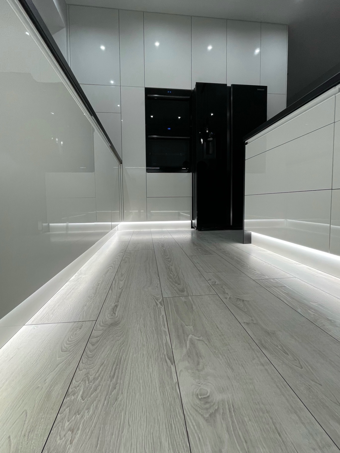 Minimalistyczna kuchnia z białymi, lakierowanymi frontami, czarnymi sprzętami AGD w zabudowie i podświetleniem LED przy podłodze z paneli imitujących drewno.