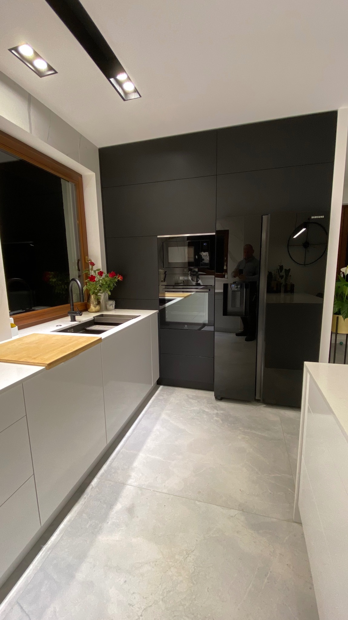 Nowoczesna kuchnia z białymi szafkami dolnymi, czarnymi szafkami górnymi, drewnianym blatem roboczym, czarną armaturą i sprzętem AGD w zabudowie, oświetlona lampami sufitowymi.