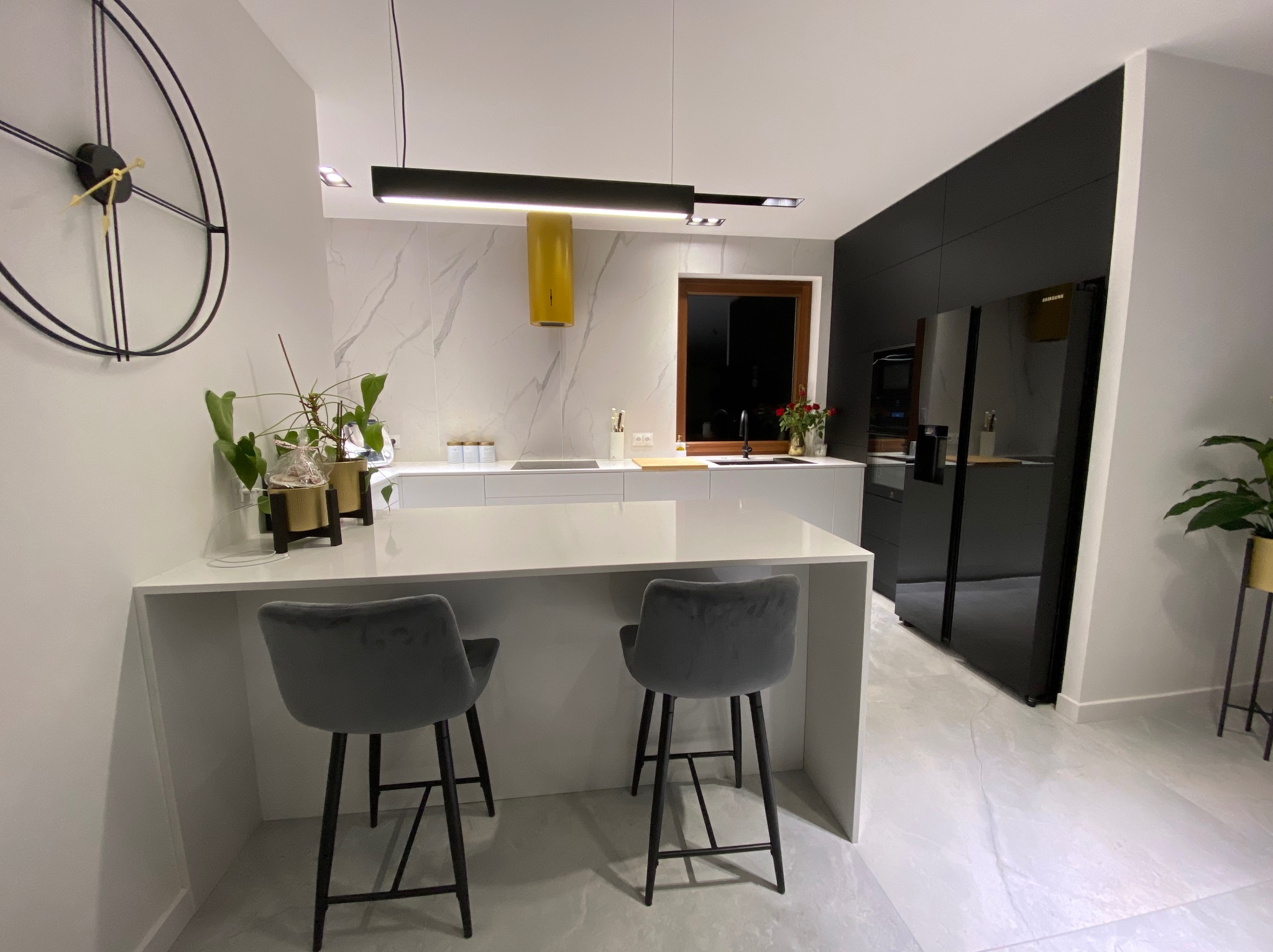 Nowoczesna kuchnia z wyspą, szarymi hokerami, czarną lodówką i żółtym okapem na tle marmurowej ściany, oświetlona podłużną lampą.