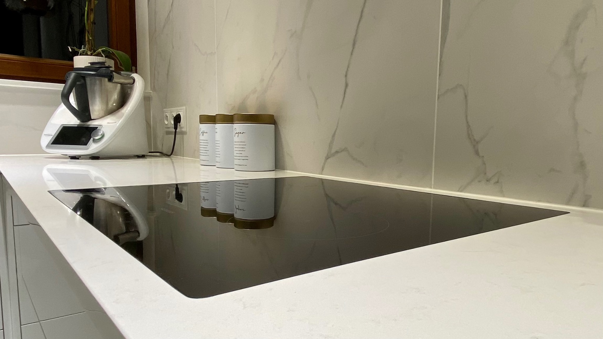 Nowoczesna kuchnia z białym blatem imitującym marmur, indukcyjną płytą grzewczą, robotem kuchennym Thermomix i zestawem pojemników na przyprawy.