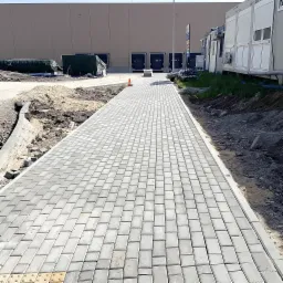 Świeżo ułożona, długa ścieżka z szarej kostki brukowej prowadząca w kierunku magazynu z rampami, widoczne prace wykończeniowe po bokach, pomarańczowy pachołek ostrzegawczy na ścieżce.