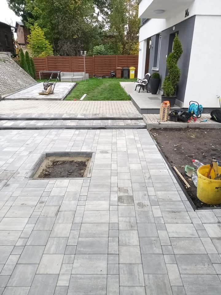 Fragment posesji z częściowo ułożoną szarą kostką brukową, widoczny otwór przygotowany pod nasadzenie rośliny, w tle trawnik, ogrodzenie i budynek.