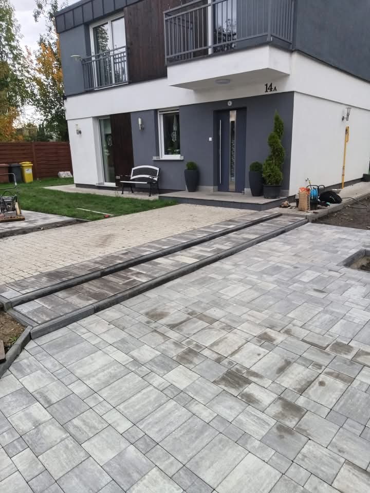 Niedawno ułożona szara kostka brukowa przed nowoczesnym domem z numerem 14A, widoczny fragment trawnika, ławka i narzędzia budowlane.