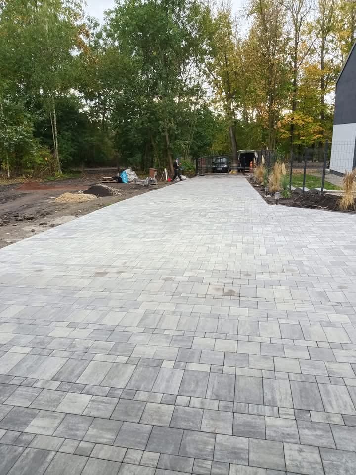 Ułożona szara kostka brukowa tworząca podjazd do posesji, widoczny fragment domu, drzewa w tle, w oddali zaparkowany samochód dostawczy i pracownik.