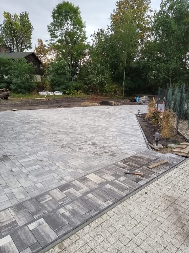 Ułożona szara kostka brukowa na podjeździe z widocznym młotkiem brukarskim i fragmentami kostki granitowej, w tle drzewa i budynek.