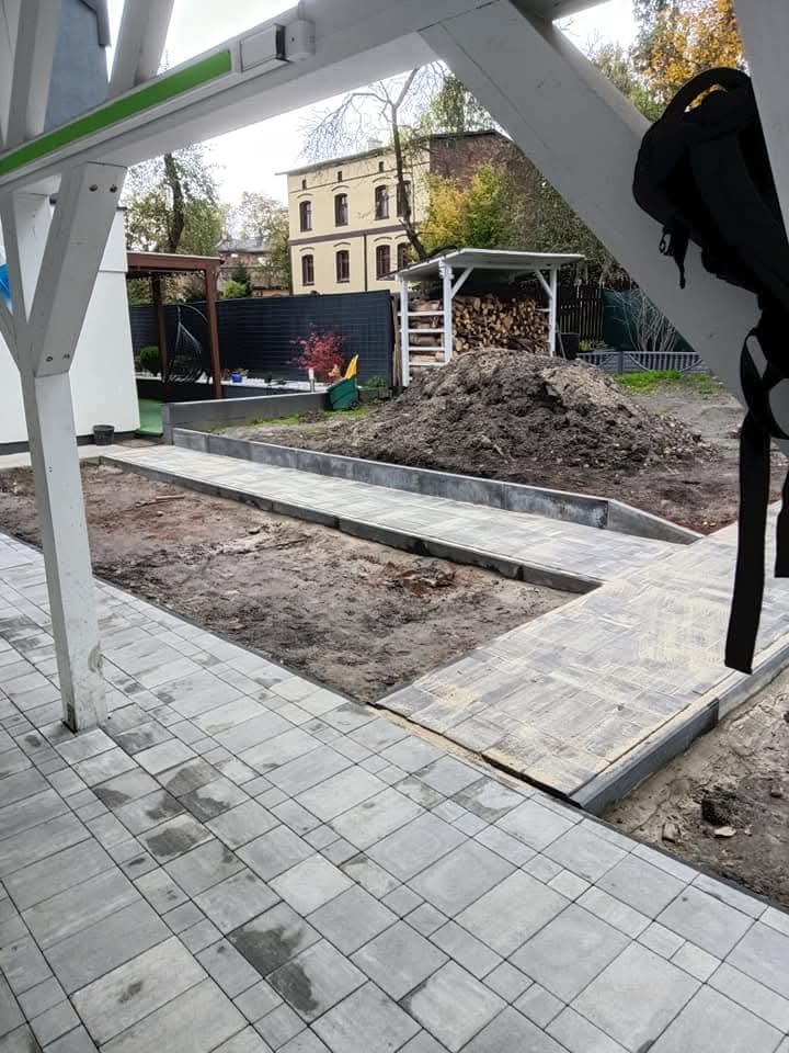 Niedokończona ścieżka z szarej kostki brukowej na posesji z widocznym stosem ziemi, drewutnią i budynkiem w tle, ujęcie zza konstrukcji altany.