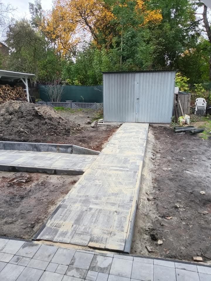 Świeżo ułożony chodnik z szarej kostki brukowej prowadzący do metalowego garażu na tle jesiennego ogrodu, widoczny stos drewna i kupka ziemi.