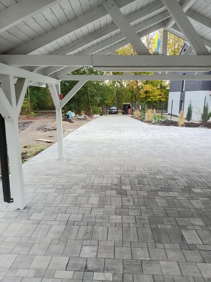 Ukończony podjazd z szarej kostki brukowej ułożonej we wzór, zadaszony białą konstrukcją drewnianą, w tle zieleń drzew i budynek mieszkalny.