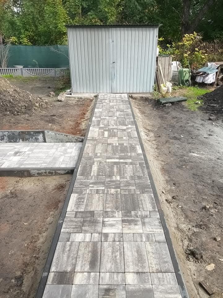 Ścieżka z szarej kostki ułożona prowadząca do metalowego garażu, widoczny fragment przygotowanego podłoża pod kolejną część nawierzchni.