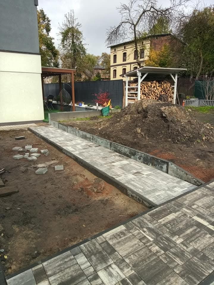 Ścieżka z szarej kostki brukowej w trakcie układania, widoczne niezakończone fragmenty, ziemia, w tle dom i sterta drewna.