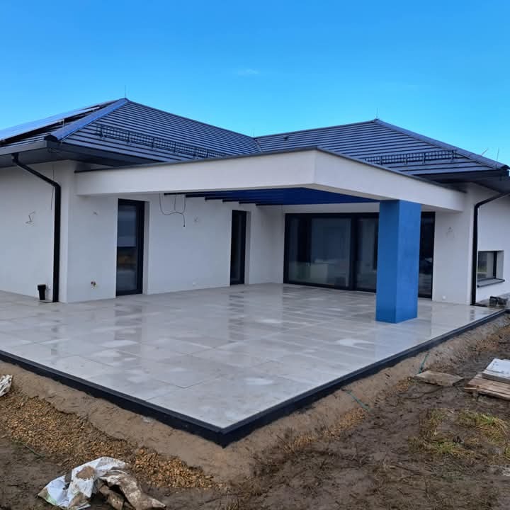 Nowoczesny taras wyłożony jasnymi płytami, obrzeża zabezpieczone czarnym profilem, z widocznym fragmentem domu z niebieskim słupem podtrzymującym zadaszenie i fragmentem niezabezpieczonego terenu.