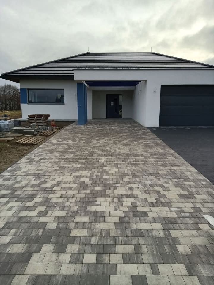 Ułożona kostka brukowa w odcieniach szarości prowadząca do nowoczesnego domu z ciemną stolarką i garażem, palety kostki w tle.